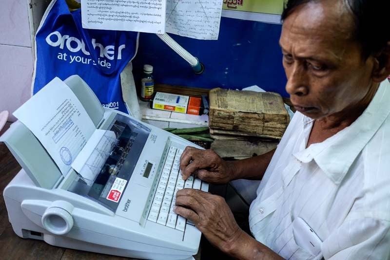 A man works on a document using an electric typewriter inside a downtown Yangon shop. While typists traditionally station themselves outdoors near courts and government offices, small indoor shops are opening in the vicinity, featuring more modern typewriters, as well as copiers and computers. (Photo and caption: Tin Htet Paing / The Irrawaddy)
