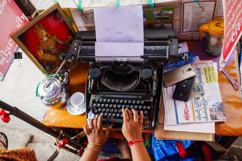 A female typist works on a document from her kiosk on lower Maha Bandoola Park Road. Like an increasing number of typists in Yangon, she relies on a typewriter professionally, but for personal purposes, she uses a smartphone. (Photo and caption: Tin Htet Paing / The Irrawaddy)