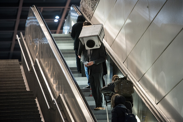 Personne portant une vidéo de surveillance en carton sur sa tête dans des escalators