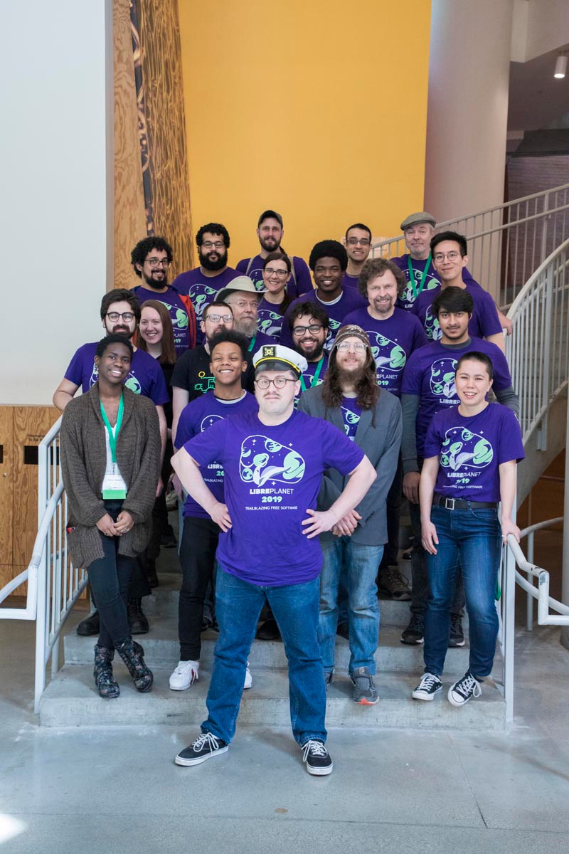 LibrePlanet volunteer captain Matt Lavallee posed in front of a staircase with a large group of LibrePlanet volunteers in matching purple LibrePlanet shirts